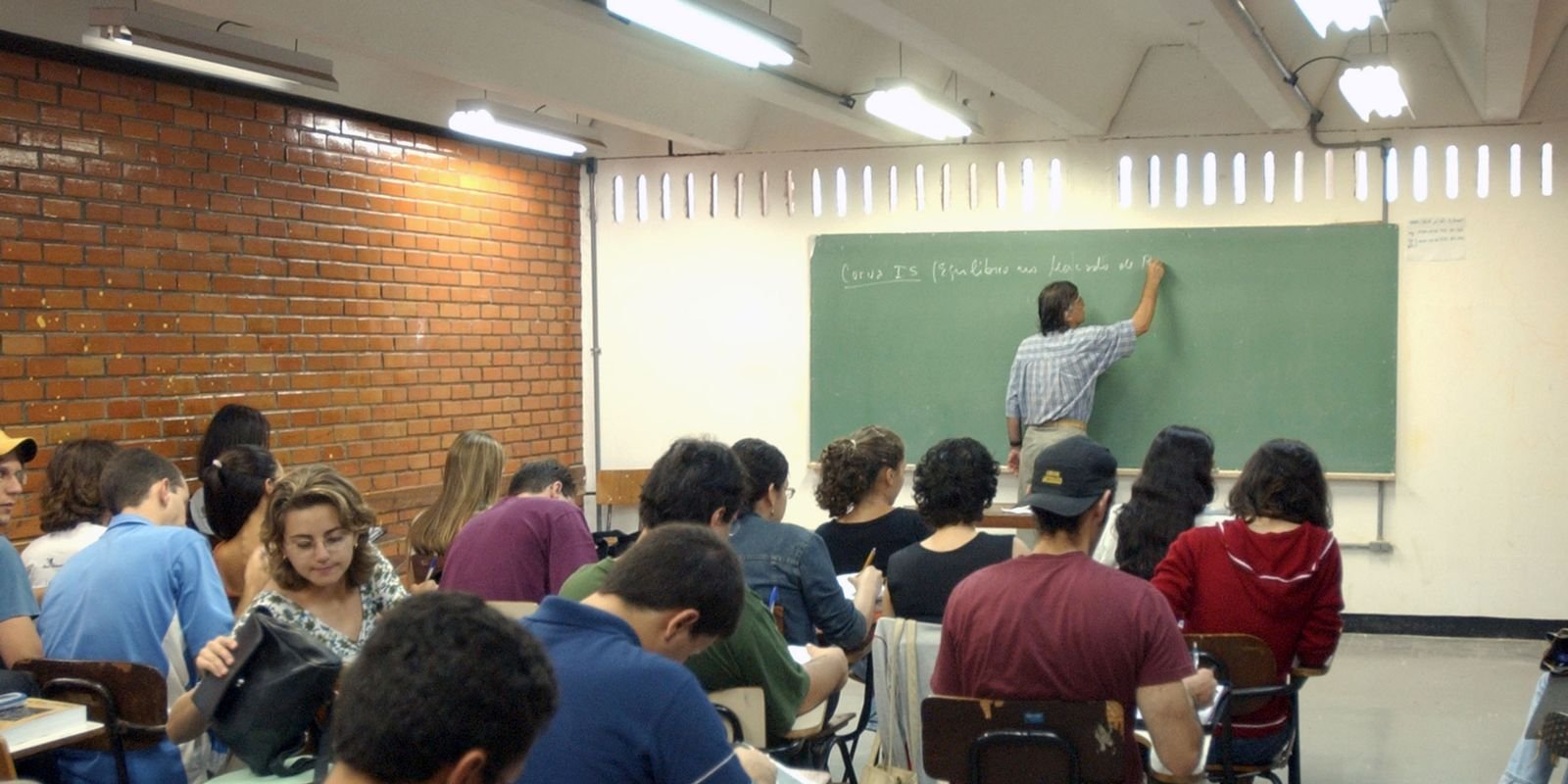 Professor escreve no quadro enquanto estudantes acompanham uma aula, em fotografia de arquivo.