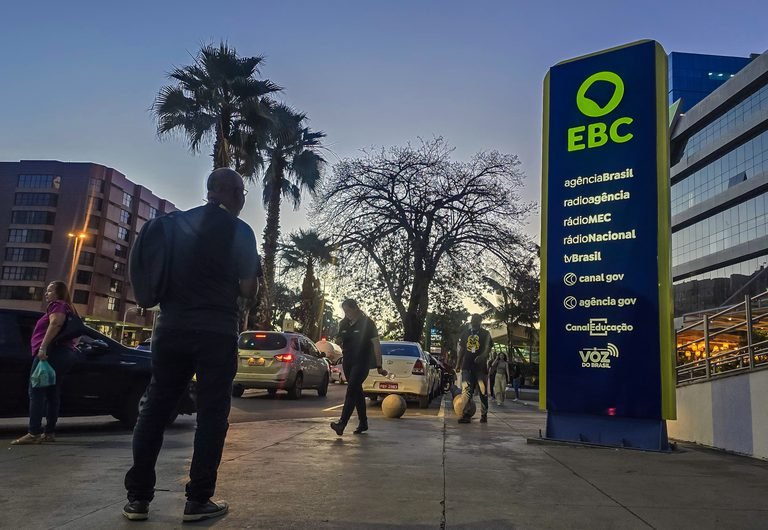 Pessoas caminham diante de prédio e placa institucional da EBC, em imagem de divulgação.