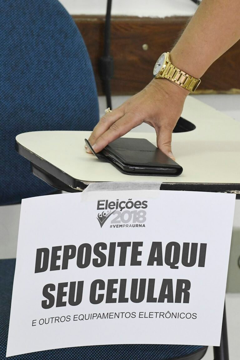 Pessoa deixa celular em caixa identificada para guardar aparelhos fora da cabine, nas eleições de 2018.