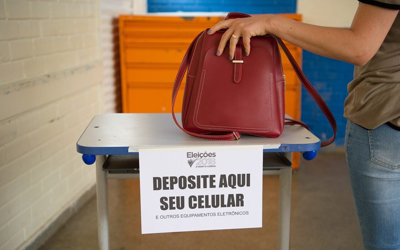 Eleitora ao lado de mesa com aviso para guardar celulares em seção eleitoral de 2018.