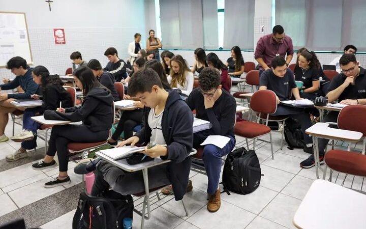 Estudantes sentados em carteiras preenchem provas em sala de aula.