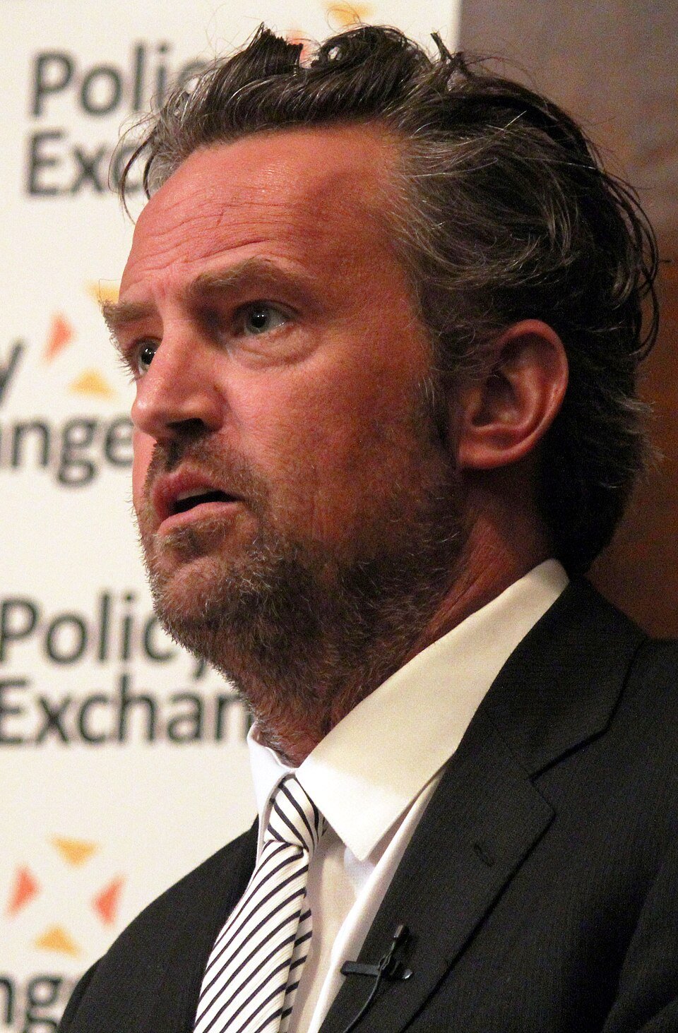 Matthew Perry em evento, vestindo paletó escuro e gravata.