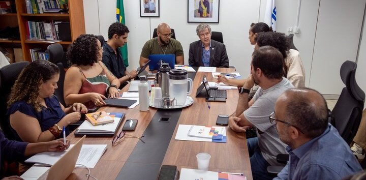 Participantes com computadores e documentos reunidos em torno de uma mesa.
