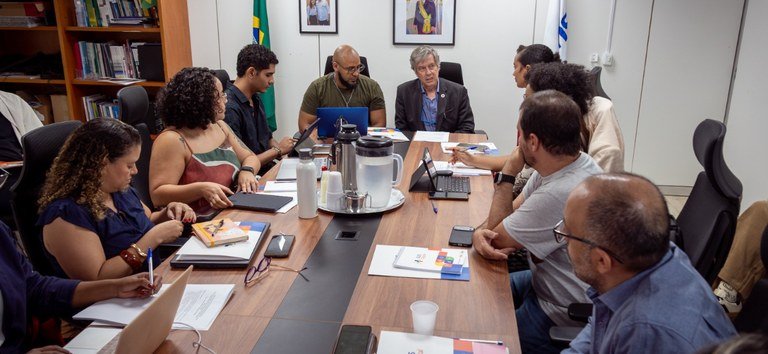 Participantes com computadores e documentos reunidos em torno de uma mesa.