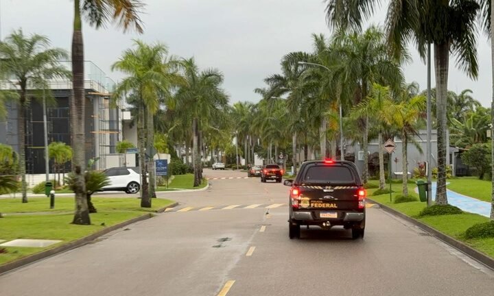 Viaturas da Polícia Federal circulam por uma rua arborizada.