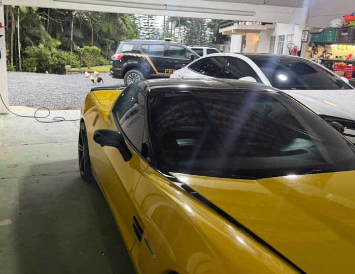 Carro esportivo amarelo em garagem, com viatura da Polícia Federal ao fundo.