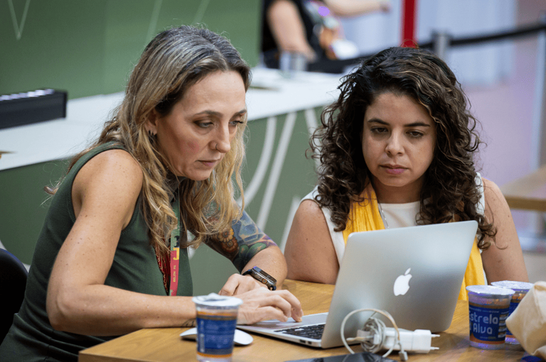 Duas mulheres consultam um computador portátil durante atividade de trabalho.